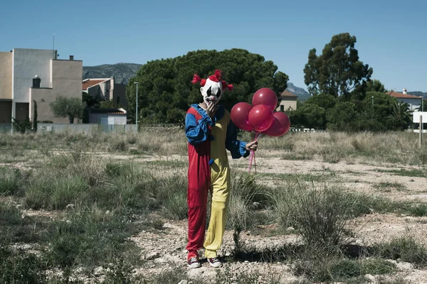 a creepy clown wearing a yellow, red and blue costume, holding a bunch of red balloons in his hand, standing in a vacant lot