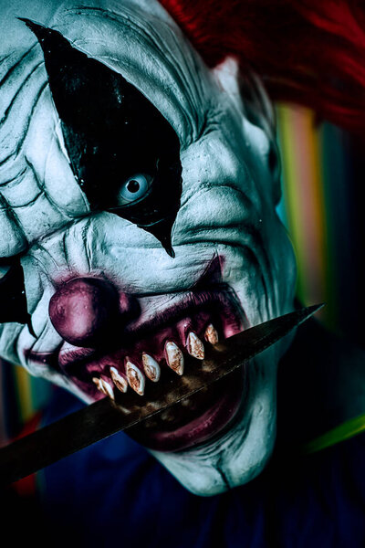 closeup of a scary evil clown, with red hair, white eyes and bloody teeth, with a big knife in his mouth staring at the observer with a threatening look