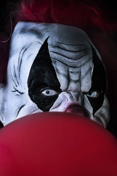一个有着红头发和白眼睛的可怕的邪恶小丑的特写 他盯着观察者 前面有一个红色气球 背景是黑色的 — 图库照片