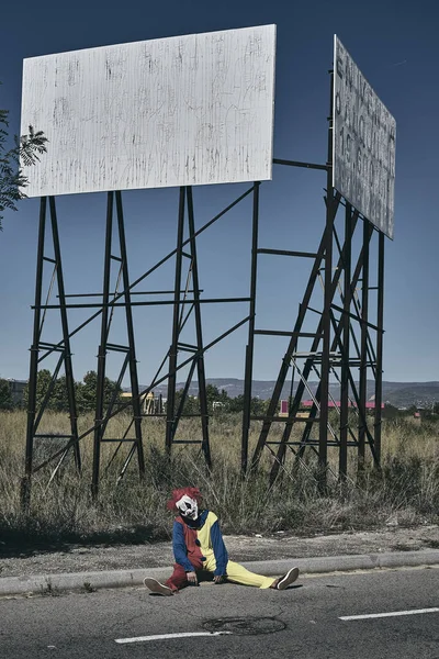 一个可怕的邪恶小丑 穿着黄色 红色和蓝色的服装 坐在一个废弃的广告牌锈迹斑斑的结构前面的乡间路上 — 图库照片