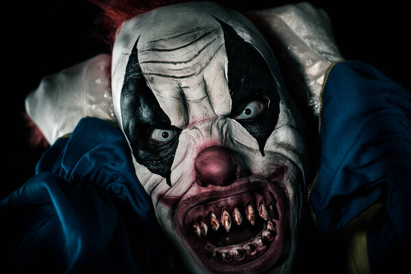 closeup of a scary evil clown with frightening white eyes and bloody teeth, pulling his red hair while staring at the observer with a threatening look