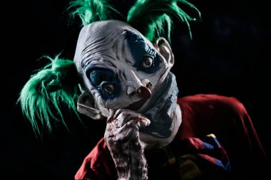 Yeşil saçlı, kirli kırmızı bir kostüm, kirli bir papyon takan, kirli ve kanlı eldivenler takan iğrenç maskesini aşağı ya da yukarı siyah bir arka plana doğru çeken rahatsız edici kötü bir palyaçonun yakın çekimi.