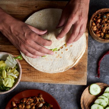 Kafkasyalı genç bir adamın soğan, kırmızı ve yeşil biber, taze marul ve çiğ salatalık gibi farklı sebzelerle pişirilmiş tavuk eti ya da burrito hazırlarken yüksek açılı görüntüsü.