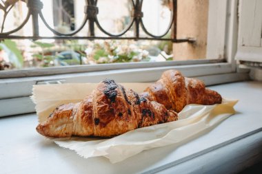İki kruvasan beyaz bir pencere pervazının üzerinde duruyor. Biri siyah çikolata lekeli, diğeri ise dışarıda bulanık çiçekli.