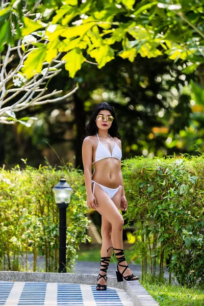 Young asian woman in the white bikini sun-tanned skin is posing near the swimming pool in a hot weather
