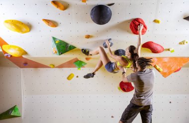 Spor tırmanış içinde kaya dağcılar. Genç kadın Bouldering sorun (rota) taşkın duvara erkek eğitmen onu güvenliğini sağlama tırmanma.
