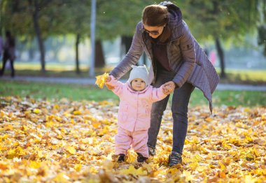 Bebek kızı destekleyici ve yardımcı olun ilk adımlarını anne. Anne ve kızı sonbahar parkta yürümek kadar kolay üzerine.