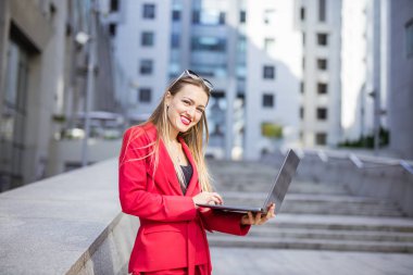 Ofis binalarının önünde dizüstü bilgisayar kullanan çekici genç bir kadın.