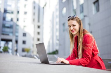 Ofis binalarının önünde dizüstü bilgisayar kullanan çekici genç bir kadın.
