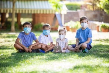 Dışarıda yüzlerinde koruyucu maskeler olan küçük çocuklar. Karantinaya alın. Çocuklar parkta çimlerin üzerinde otururken güvenlik maskesi takıyorlar. Coronavirus önleme.