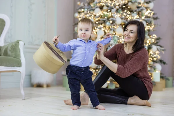 クリスマスツリーの近くに赤ちゃんの孫と遊ぶ魅力的な成熟した女性 男の子はギフトボックスを持っている — ストック写真