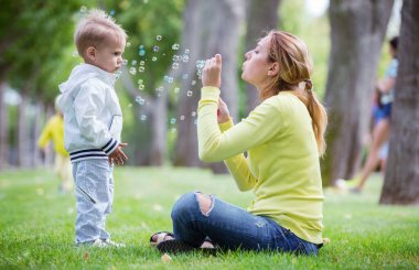 Anne dışarıda sabun köpüğü üflüyor, küçük oğlu da onu izliyor. Aile parkta eğleniyor..
