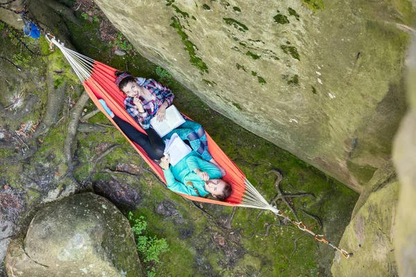 Young women reading books and eating chocolate while relaxing in hammock near cliff, view from above