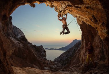Rock climbing at sunset. Young man climbing route in cave, female partner belaying him. Cave shaped  clipart