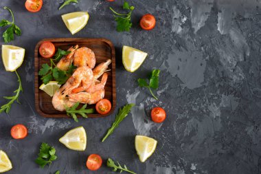 Bir kare ahşap tabak karides vardır. Çanak maydanoz, limon, rucola ve kiraz domates dağılmış. Siyah gri beton arka plan. Metin için yer kopyalayın.