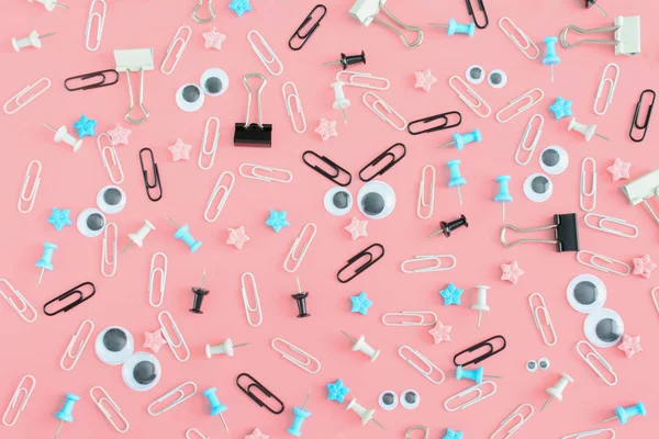 Evil smiley. Face with puppet eyes and hair from clips on a pink background. The stationery is chaotically scattered. An unusual background of clips and clerical buttons. Beautiful picture from the top.