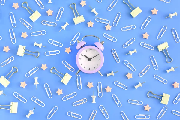 White paper clips, clerical buttons and small pink stars are in disorder on a blue background. In the center lies a small beautiful pink alarm clock. Stationery chaotically scattered. Beautiful photo from the top.