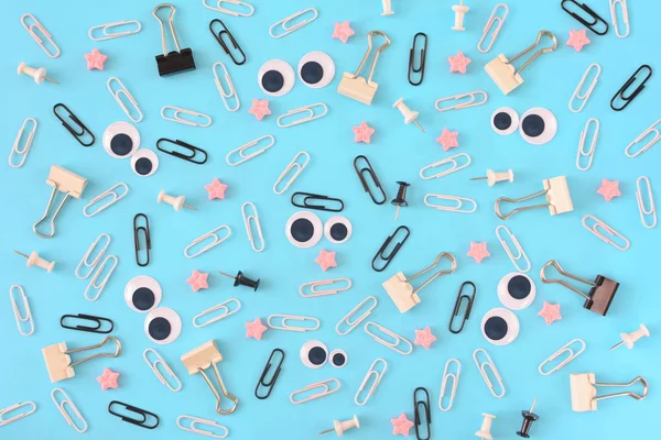 White and black paper clips, pink stars and stationery are scattered chaotically on a blue background. Funny smiley from clips in the center. Bright photo from the top.