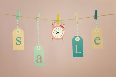 İpi asmak bir hafif pembe üzerinde Satılık etiketlerini etiketli. Küçük pembe çalar saat merkezidir. Kavram, satış için zaman.