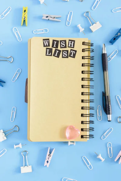 No centro encontra-se um caderno aberto sobre molas com páginas castanhas. Papelaria em desordem em um fundo azul. Lista de desejos de inscrição. Faz asneira. . — Fotografia de Stock