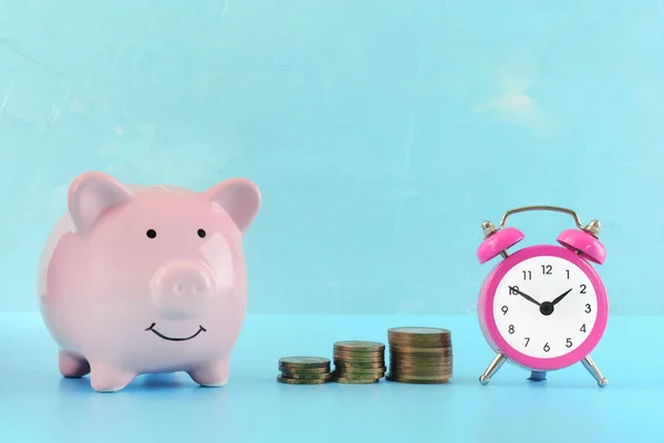 Un pequeño despertador rosa, tres pilas de monedas y una alcancía sobre un fondo azul claro. El símbolo de la acumulación de dinero. Brillante foto . —  Fotos de Stock