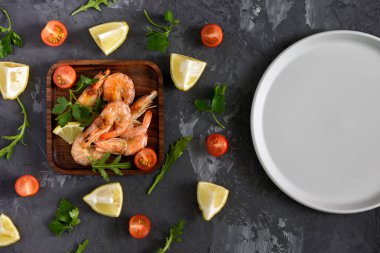 Ahşap bir tabak karides vardır. Sonraki siyah dokulu bir arka plan üzerinde otlar, baharatlar ve limon dağınık. Boş bir gri levha.