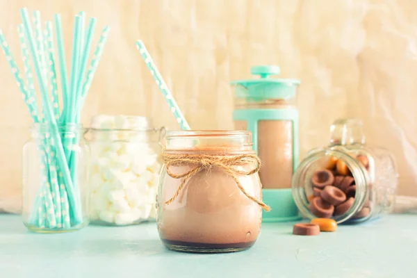 El cacao caliente se vierte en el frasco. En el fondo cafetera, malvaviscos y dulces de chocolate. El fondo en el bokeh . — Foto de Stock