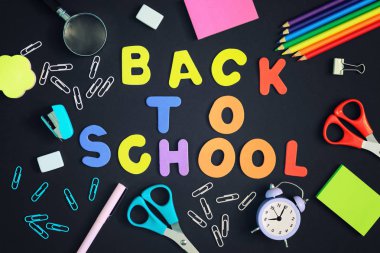 Siyah bir arka plan üzerinde ataç, bir dizüstü bilgisayar, bir çalar saat ve okul malzemeleri vardır. Back to School renkli harflerle yazıt ortasında. Parlak fotoğraf.