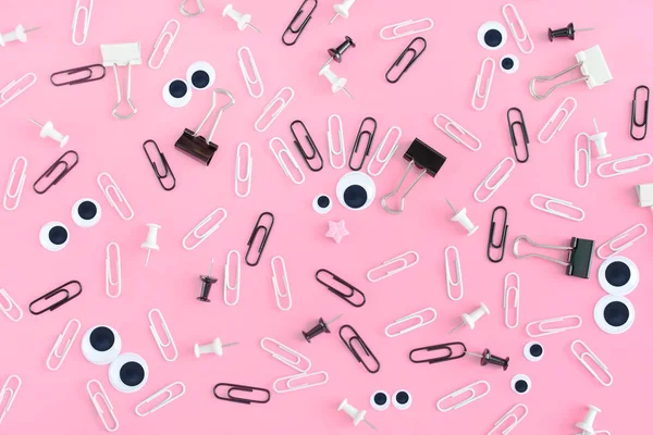 Crazy smiley. Face with puppet eyes and hair from clips on a pink background. The stationery is chaotically scattered. An unusual background of clips and clerical buttons.Photo from the top.