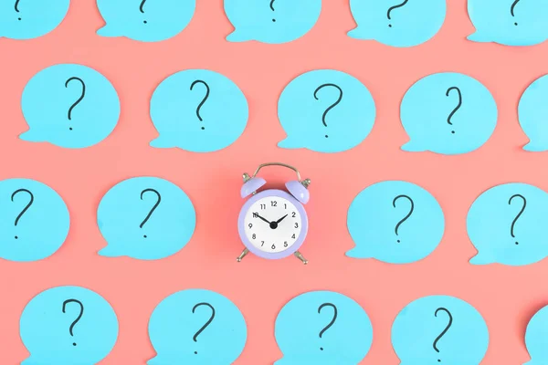 Blue stickers with question marks on a pink background. In the center is a small pink alarm clock. Concept, a matter of time. Beautiful bright photo from the top.