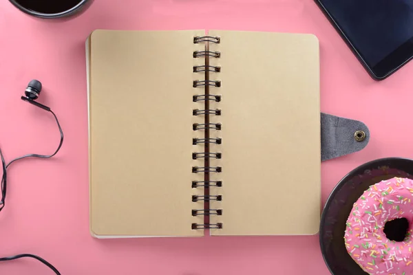 A notebook on springs with brown pages on pink background with donut and headphones.