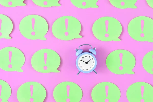Bright yellow stickers with exclamation marks on a pink background. In the center is a small lilac alarm clock. Concept, attention time. Beautiful bright photo from the top.