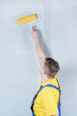 Mavi tulumlu ve sarı tişörtlü bir adam silindirle çalışıyor. Ev ressamı duvarları harçla kaplar.