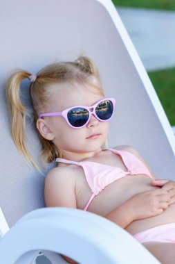 Sarı saçlı küçük bir kız güneş gözlüğüyle güneşlebe. Çocuk tatilde, şezlongda yatıyor..