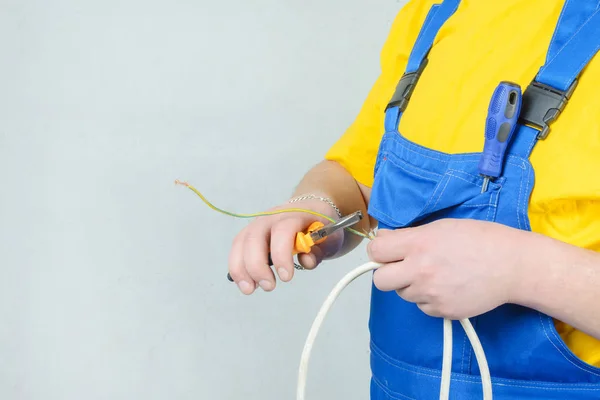 Um homem de fato-macaco azul e uma T-shirt amarela, a segurar alicates e fios. Obras de eletricista . — Fotografia de Stock
