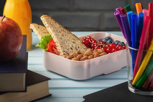 Healthy food for school lunch. On the table among the textbooks is a school lunch, in a box are almonds, red currants and blueberries. Nuts, berries and a sandwich for a childs snack — Stock Photo, Image