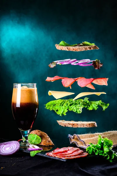 Sandwich met vallende ingrediënten in de lucht en donker bier in een — Stockfoto