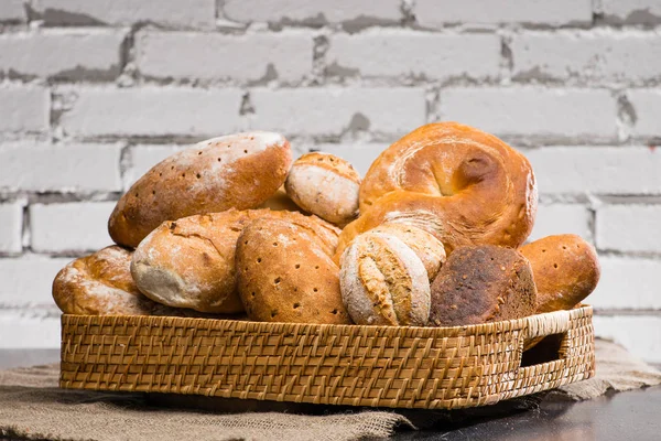 Various types of fresh bread in a basket on a table, brick backg — Stock Photo, Image