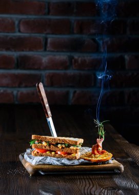 Lezzetli doğal ve diyet sandviçi, bir bara saplanmış bıçak ve biberiye dumanı, tuğla duvardaki yazı için fotokopi yeri, avakado, domates ve roketli pastırma sandviçi ahşap arka planda.