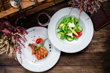 Restoran masası iki ana yemek için hazırlandı. Biftek mutfağı, kızarmış patates, kaliteli şarap kadehi. Ahşap bir masada iki tabak - biftek ve salata. Öğle yemeği için yemek.