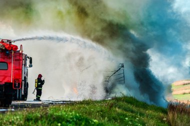 İtfaiyeci kazadan sonra yanan bir arabayı söndürür. İtfaiyeci su ve yangın söndürücü kullanır. İtfaiyeci yangın söndürücü kullanır.