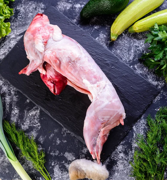 Raw whole rabbit with fresh vegetables for tasty rabbit cooking. Preparation of hunting stew on dark background, top raw rabbit carcass meat for sale