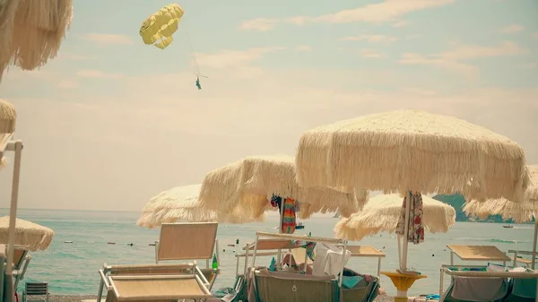 Waving beach umbrellas and distand parasailing parachute at sea — Stock Photo, Image