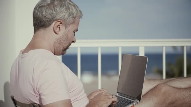 Красивый седой мужчина работает на ноутбуке на террасе с видом на море под ярким солнцем — стоковое видео