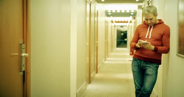Γκρι μαλλιά νεαρός άνδρας στο casual ρούχα χρησιμοποιεί το smartphone του στο διάδρομο ξενοδοχείου — Αρχείο Βίντεο