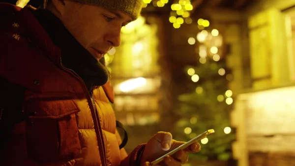 Schöner junger Mann in roter Jacke nutzt abends sein Smartphone gegen Weihnachtsbeleuchtung — Stockfoto