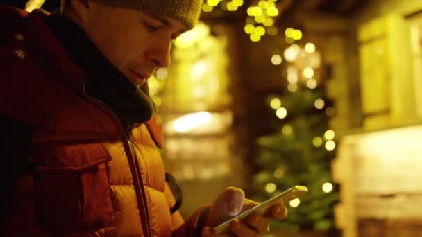 Pohledný mladý muž v červeném kabátku používá své smartphone večer proti vánočním osvětlením. Střela na červeném fotoaparátu — Stock video