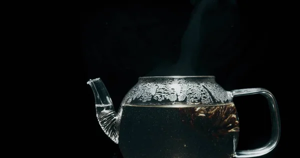 Steaming hot tea in a glass teapot against black background