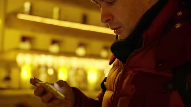 Красивый молодой человек в красной куртке смотрит фотографии на своем смартфоне вечером — стоковое видео