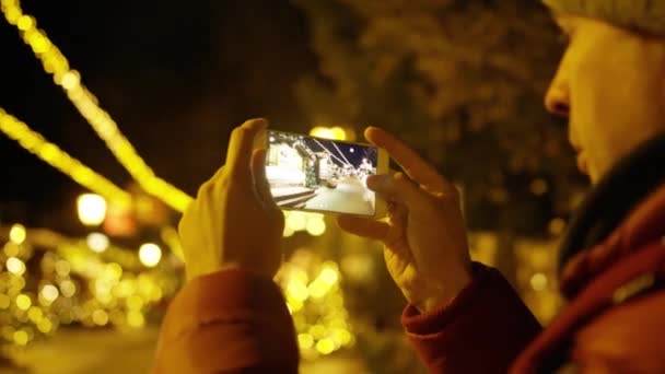 Ein junger Mann in roter Jacke macht abends mit dem Smartphone Fotos vor weihnachtlichem Hintergrund. Aufnahme mit roter Kamera — Stockvideo